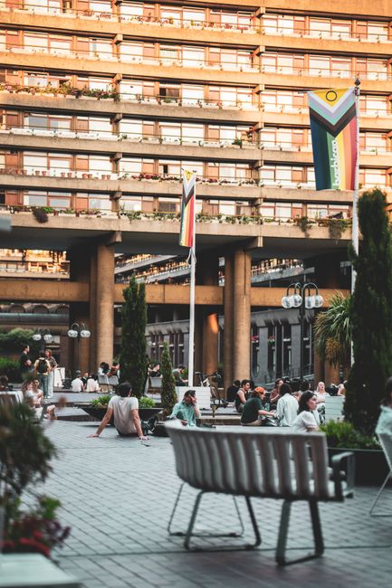 A large urban building complex featuring Brutalist architectural elements, including concrete facades with exposed textures and geometric shapes. The foreground shows a paved outdoor area with concret