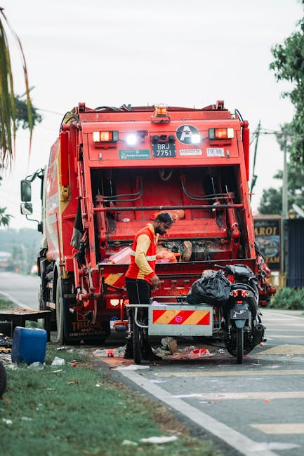 A worker in a high-visibility yellow and red jacket stands beside a large red rubbish collection truck on a street, actively clearing waste. The truck's rear mechanism is open, revealing internal comp