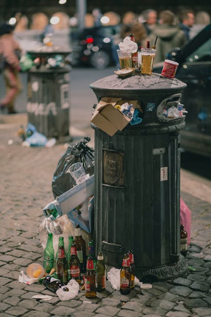 A black cylindrical rubbish bin with a partially open white plastic lid, positioned against a beige stone wall. The bin is overflowing with various types of trash, including foil food containers, pape